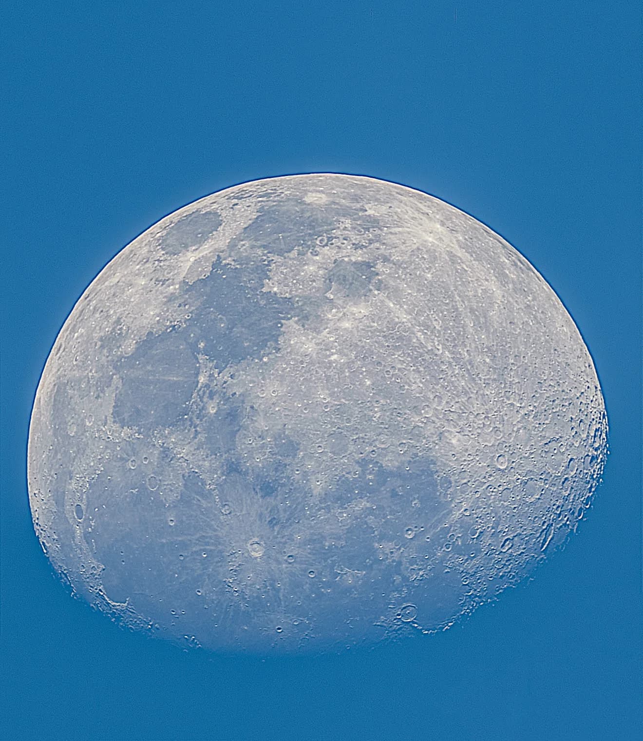 A waxing gibbous moon captured against a bright blue daytime sky, highlighting the dark lunar maria.